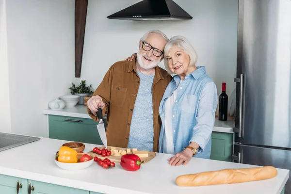 Sorrindo Casal Idosos Abraçando Perto Mesa Com Queijo Baguete Legumes — Fotografia de Stock