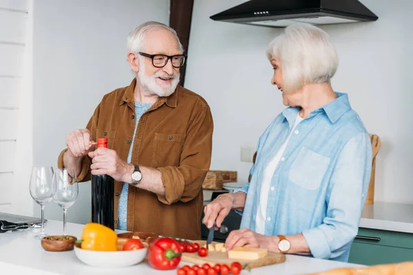 Happy Elderly Husband Looking Wife Cutting Cheese While Opening Bottle — Stock Photo, Image