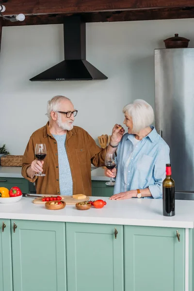 Smiling Elderly Husband Wine Glass Feeding Wife Piece Cheese Kitchen — Stock Photo, Image