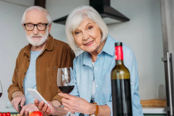 Smiling Senior Woman Smartphone Wine Glass Looking Camera Husband Blurred — Stock Photo, Image