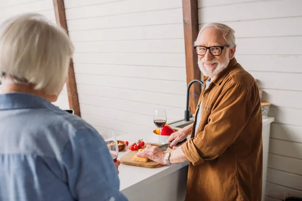 Happy Senior Man Looking Camera While Cutting Cheese Blurred Woman — Stock Photo, Image