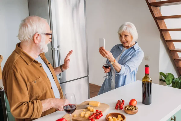 happy elderly wife with wine glass and smartphone taking photo of husband cooking dinner in kitchen