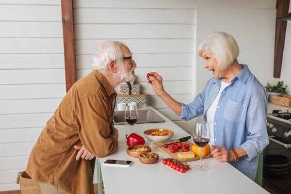 боковой вид счастливой старшей жены, кормящей мужа помидорами черри возле стола с едой, бокалами для вина и смартфоном на кухне