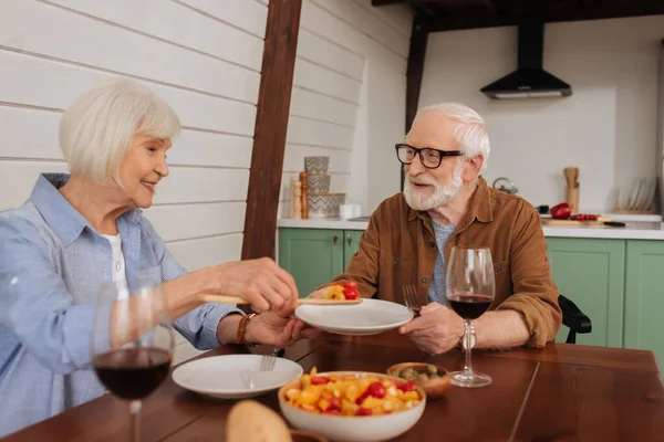 笑容满面的年长妻子 配上意大利面 为丈夫端上沙拉 晚餐时前景朦胧 — 图库照片