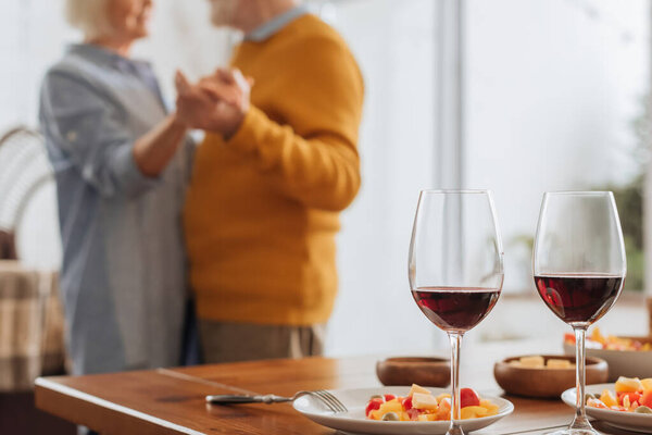 cropped view of elderly couple dancing near table with wine glasses and vegetarian salad on plates on blurred background