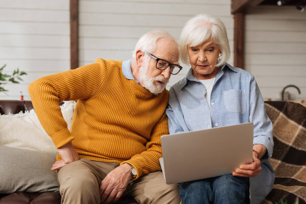 senior husband with hand on hip looking at laptop near wife on couch on blurred background