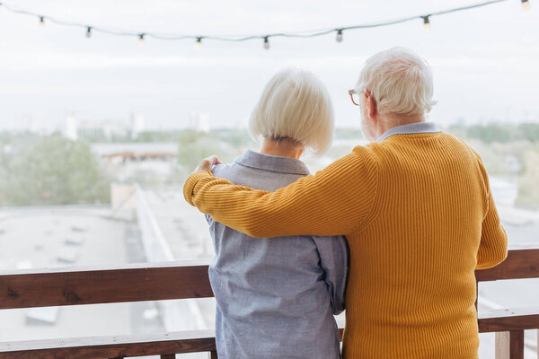 back view of senior husband hugging wife on terrace on blurred background