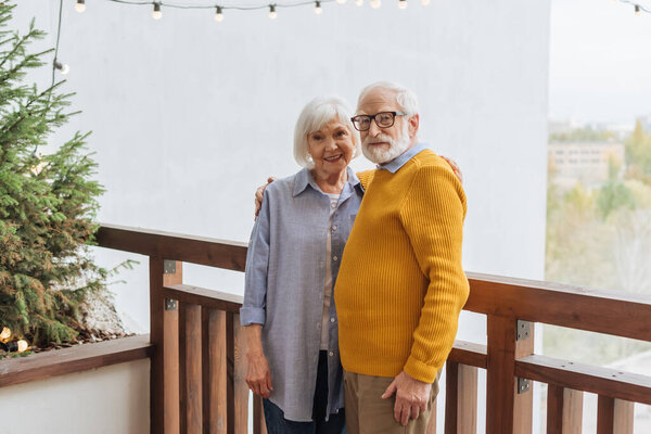 smiling senior couple looking at camera while hugging on terrace near pine tree on blurred background