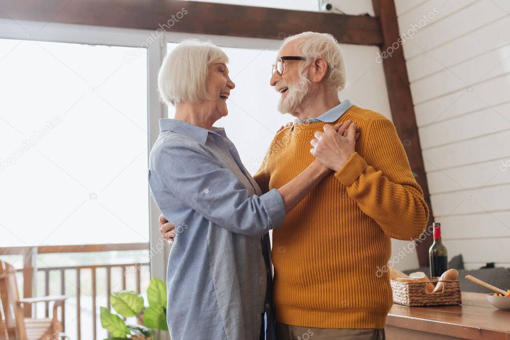 happy elderly couple laughing while dancing at home on blurred background