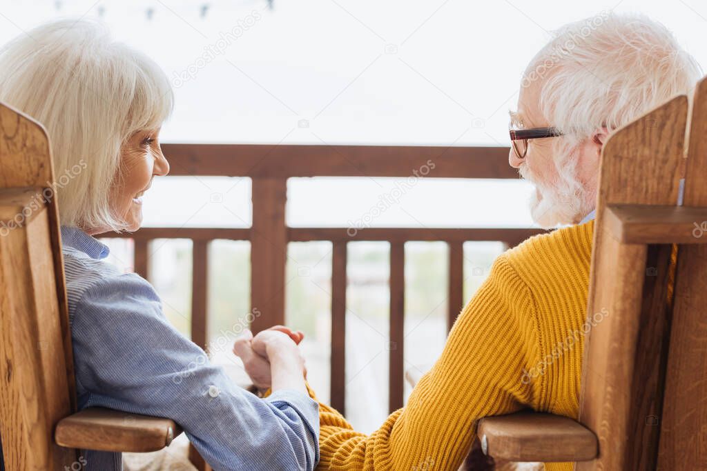 happy senior wife laughing while holding hands with husband on terrace on blurred background