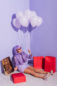 junge Frau im Puppenstil sitzend mit Einkaufstaschen und Luftballons auf violettem Hintergrund gekleidet