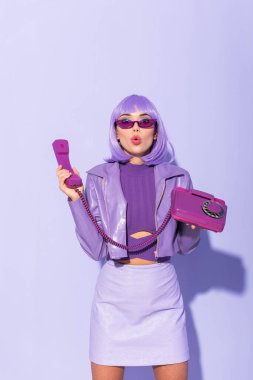 Şok olmuş, bebek gibi giyinmiş, menekşe renkli arka planda retro telefonu olan genç bir kadın.