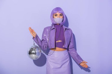 Mor renkli arka planda disko topu olan tıbbi maske takan bebek gibi giyinmiş genç bir kadın.