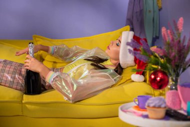 Sarı kanepede şampanyayla bebek gibi davranan Noel Baba şapkalı genç bir kadın.