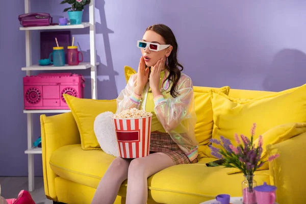 surprised young woman posing as doll with popcorn in 3d glasses on yellow couch