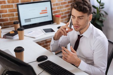 İş adamı bilgisayarın yanında akıllı telefonla konuşuyor ve ön planda bulanık bir kahve var. 