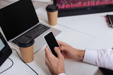 İş adamının kapalı arka planda kapalı kahve ve bilgisayarların yanında boş ekranlı akıllı telefon kullanması görüntüsü 