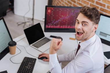 Excited businessman looking at camera while showing yeah gesture and checking financial courses on computer on blurred background  clipart
