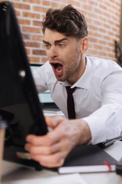 Çığlık atan iş adamı ofisteki bulanık ön planda bilgisayar monitörü tutuyor. 