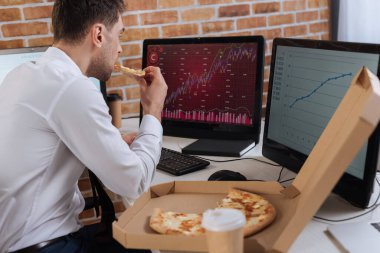 Bilgisayarların yanında pizza yiyen iş adamı. Arkaplanda grafikler var. 