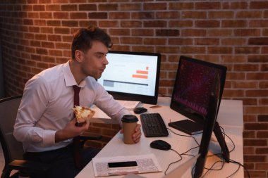 Ofisteki bilgisayar stoklarını kontrol ederken pizzayı paket kahve ile yiyen bir işadamı. 