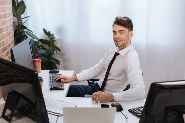 Smiling Businessman Looking Camera Computers Takeaway Coffee Newspaper Blurred Foreground — Stock Photo, Image