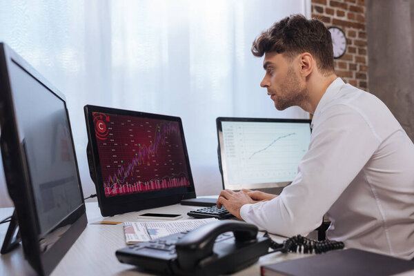 businessman using computer with graphs of finance market on monitors near telephone on blurred foreground 