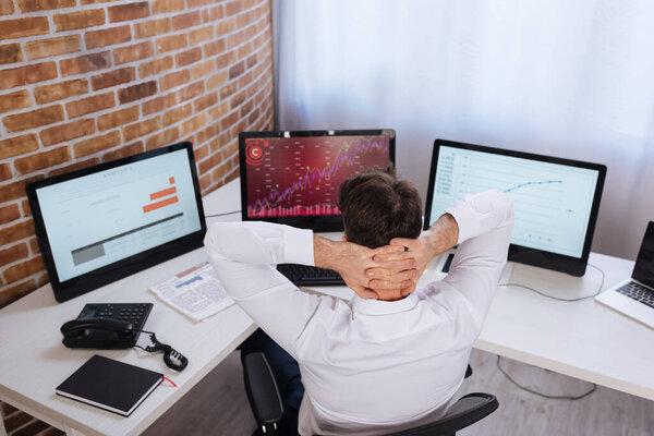 Back view of businessman sitting near computers with charts and newspaper on blurred background 