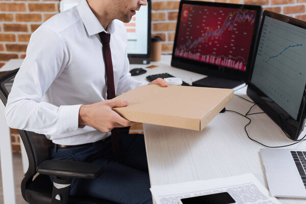 Cropped view of businessman holding pizza box near computers on blurred background in office 