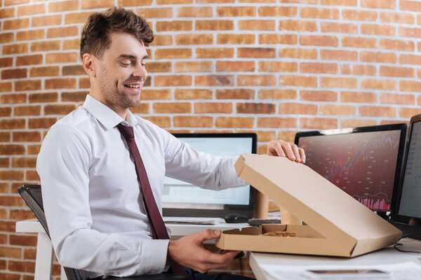 Smiling businessman opening pizza box near computers on blurred background 