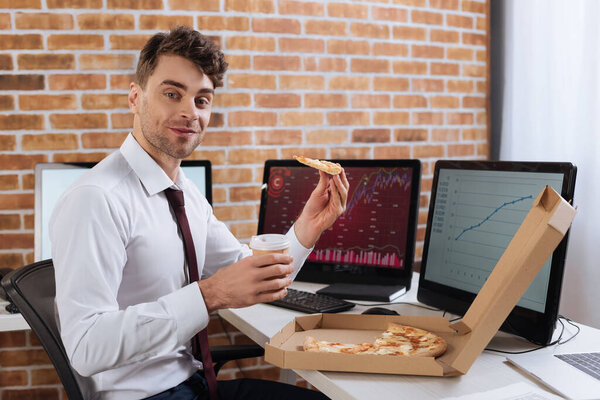 Smiling businessman holding pizza and takeaway coffee near computers on blurred background 