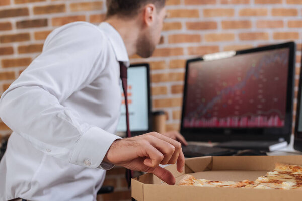 Businessman taking tasty pizza in box near computers on blurred background in office 