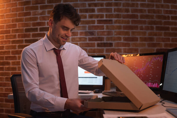Smiling businessman opening pizza box near computers on blurred background at evening 