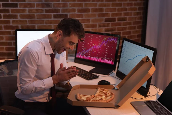 Возбужденный Бизнесмен Смотрит Вкусную Пиццу Рядом Компьютерами Графиками Размытом Фоне — стоковое фото