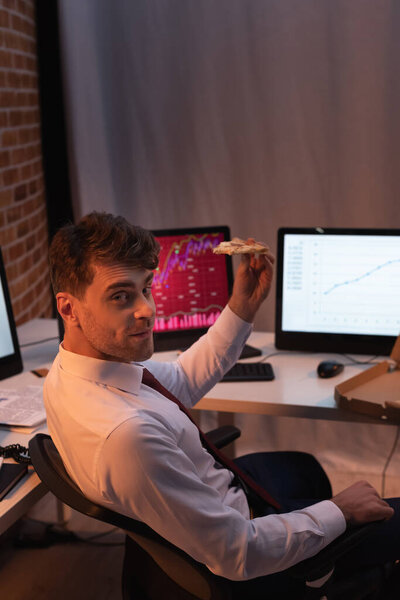 Businessman looking at camera while holding pizza near computers on blurred background 