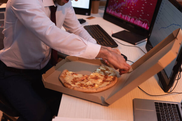 Cropped view of businessman taking tasty pizza near laptop and computers on blurred background 