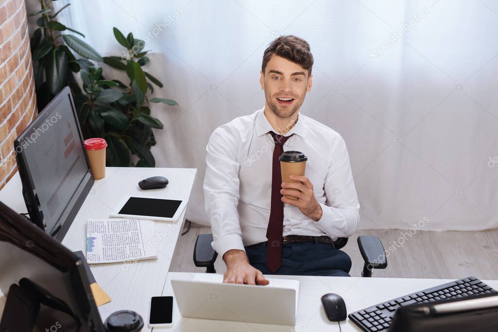 Smiling businessman holding coffee to go near devices and newspaper in office 