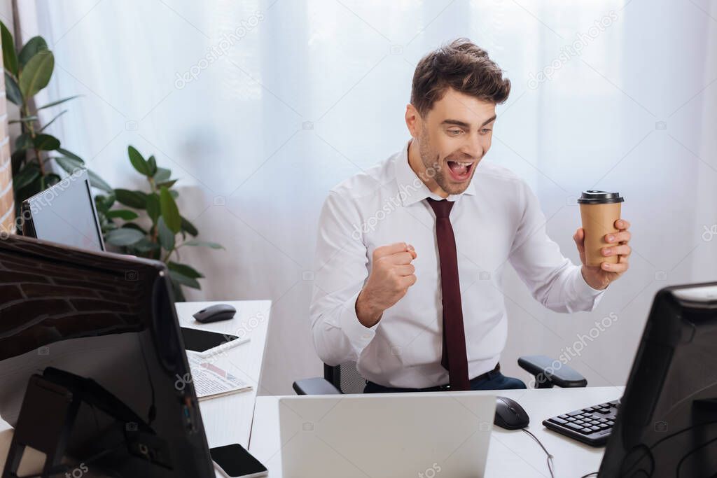 Cheerful businessman showing yes gesture while holding coffee to go and using computers on blurred foreground in office 
