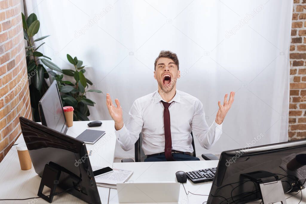 Crazy businessman screaming near devices and newspaper on blurred foreground on table 