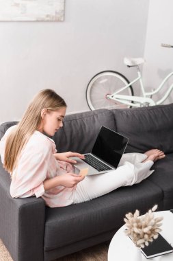 Evde e-alışveriş yaparken bilgisayarın yanında boş ekranlı kredi kartı tutan genç bir kadın.