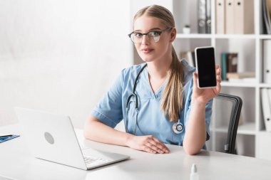 Gözlüklü veteriner masasında dizüstü bilgisayarının yanında boş ekranlı akıllı telefon tutuyor.