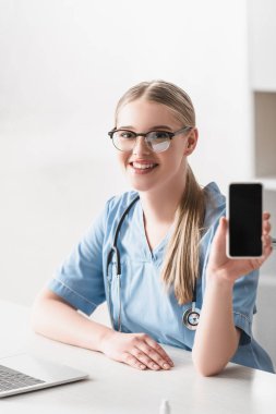 Gözlüklü neşeli veteriner masasında dizüstü bilgisayarının yanında boş ekranlı akıllı telefon tutuyor.