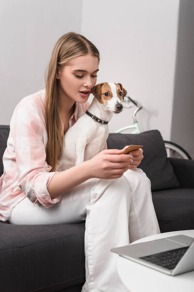 young woman holding credit card near dog and laptop on coffee table