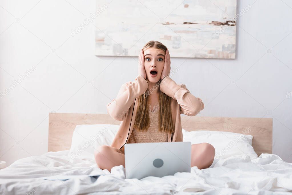 shocked freelancer in satin robe looking at camera near laptop in bedroom