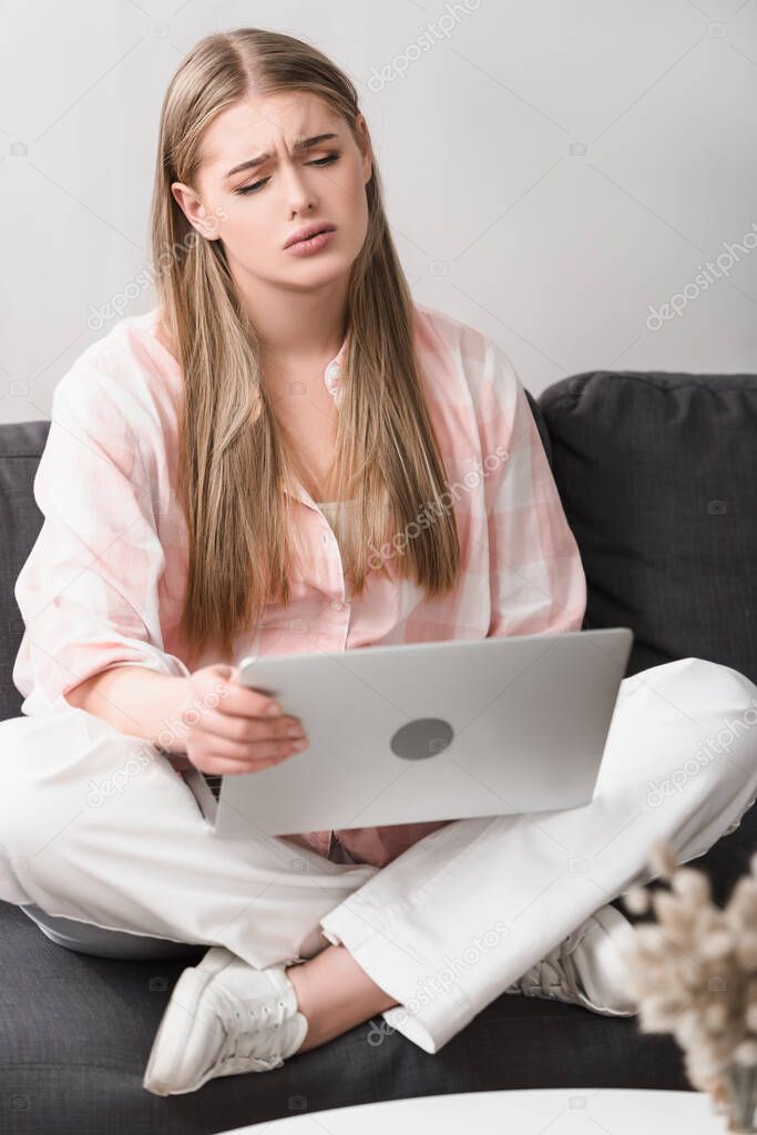 frustrated young freelancer sitting with crossed legs on couch and using laptop