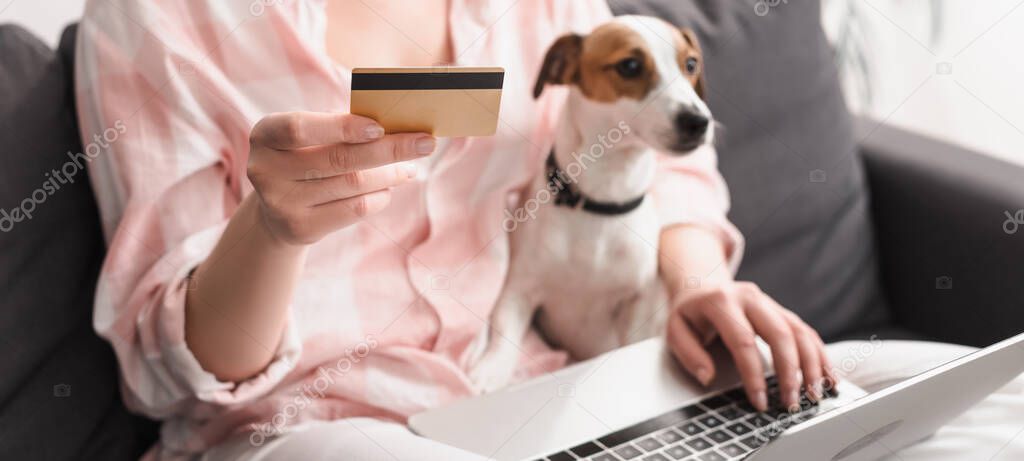 partial view of young woman holding credit card near dog and laptop while online shopping at home, banner