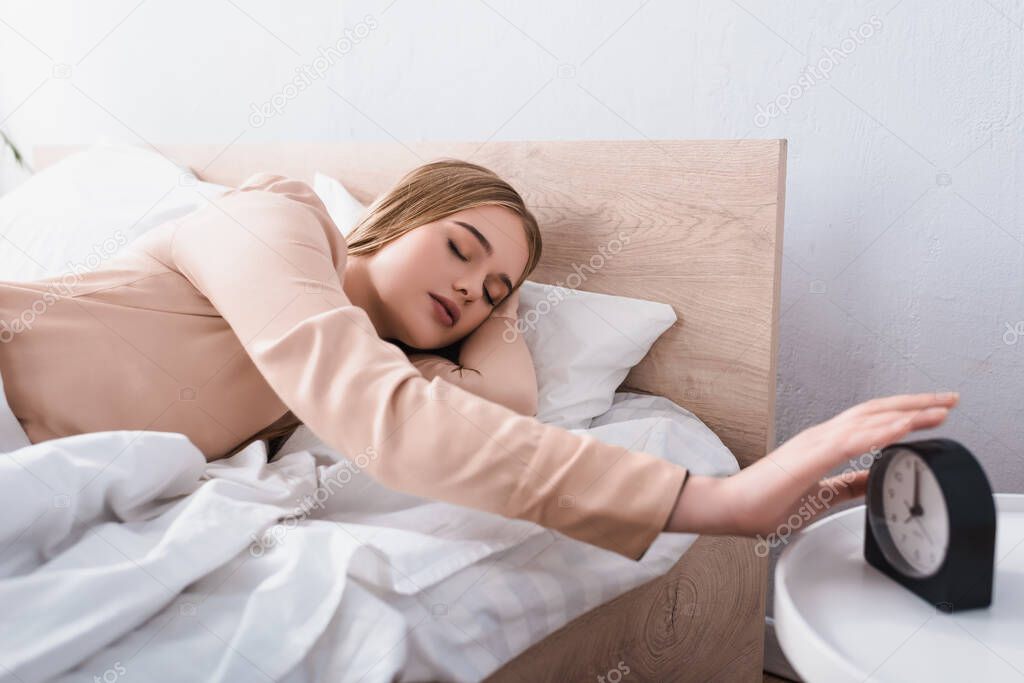 sleepy woman reaching alarm clock on bedside table 