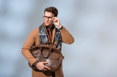 Kışlık montlu şık bir adam kahverengi evrak çantasını gri üzerinde tutarken gözlüklerini ayarlıyor.