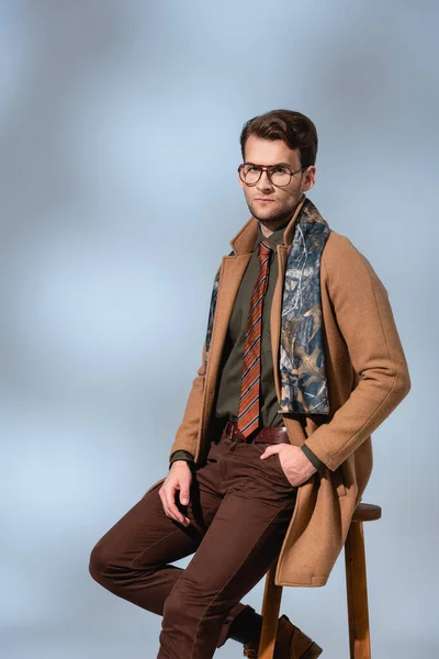 stylish man in winter outfit and glasses sitting on wooden chair with hand in pocket on grey