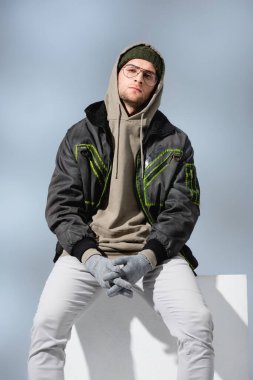 young trendy man in hat and anorak sitting on white cube with clenched hands in gloves on grey  clipart